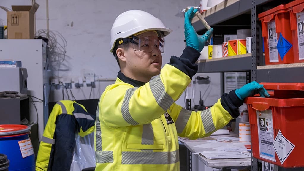 En kjemiker med gult synlighetstøy og hjelm sjekker farlig avfall. Foto.