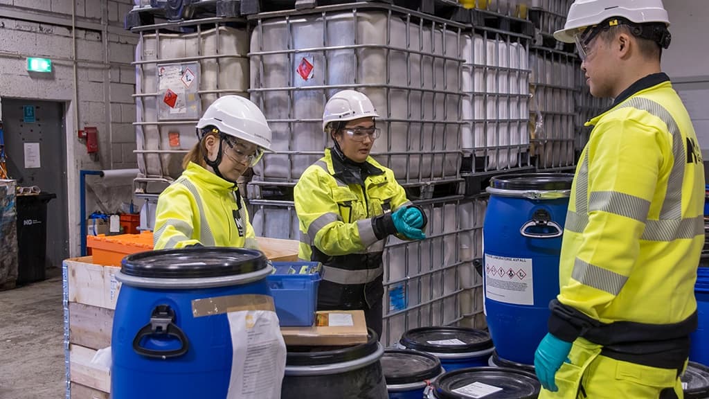Tre kjemikere med gult synlighetstøy og hjelm sjekker farlig avfall. Foto.